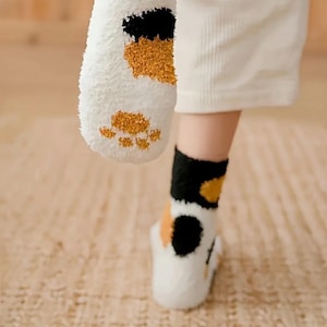 May include: White, fluffy socks with a cat paw design. The socks feature black, orange, and tan patches on the top and paw prints on the bottom. The socks are worn with white pants.