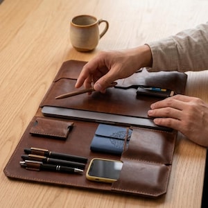 Könnte beinhalten: Eine Schreibtischunterlage aus braunem Leder mit mehreren Fächern und Taschen. Sie enthält einen Laptop, drei schwarze Stifte, ein kleines blaues Notizbuch mit kreisförmigem Muster, ein Smartphone und einen kleinen Kartenhalter auf einer flachen Oberfläche.