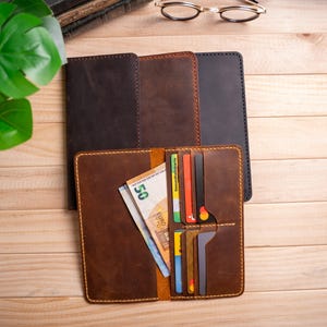 Puede incluir: Una colección de carteras largas de cuero en marrón oscuro, marrón medio y negro. Una cartera abierta de color marrón medio muestra varias ranuras para tarjetas con diversas tarjetas de crédito y un billete de cincuenta euros en el compartimento.