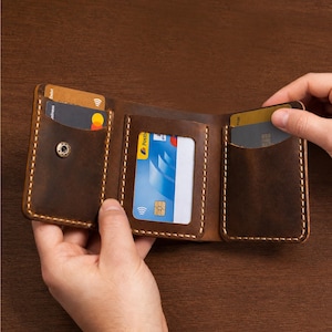 May include: A tri-fold brown leather wallet featuring visible light-coloured stitching, a metal snap button closure, and three interior card slots, including one clear window slot. The wallet is shown open with multiple credit cards inserted.
