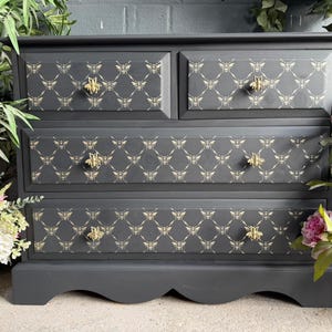 May include: A dark grey chest of drawers with four drawers. The drawer fronts have a repeating bee pattern in a lighter shade. Gold-coloured bee-shaped knobs are on each drawer. A stylish storage unit for any room.