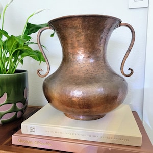 May include: A large, hammered copper vase with a wide, flared rim and two curved, scroll-shaped handles on opposite sides. The metal surface features a distinct dimpled texture and a warm, metallic brown finish with subtle variations in tone.