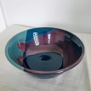 May include: A handmade ceramic bowl with a glossy finish. The bowl features a color palette of teal, deep blue, and maroon. The interior is a dark, glossy blue. The bowl is a decorative piece for serving or display.