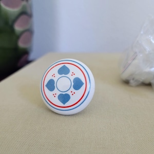 May include: A round white ceramic cabinet knob featuring a central white circle surrounded by four blue heart-shaped motifs and small red dots. The design is framed by concentric red and blue circular lines, creating a symmetrical pattern.