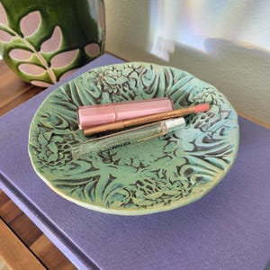May include: A round, light green ceramic dish with a textured, embossed floral pattern. Inside the dish are three items: a rectangular light pink lip product, a thin metallic gold lip liner, and a clear glass perfume vial labeled REPLICA.