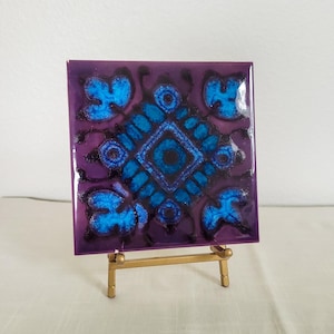 May include: Square ceramic tile with a purple background and a blue and black floral design. The tile is displayed on a small gold-colored easel. The tile's design features a central diamond shape surrounded by stylized floral elements.