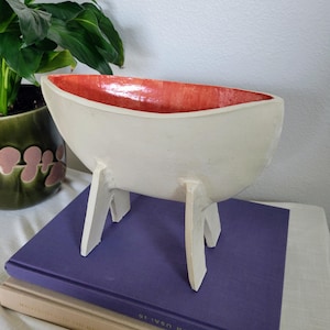 May include: A white ceramic bowl with a red interior, supported by three white legs. The bowl is placed on a stack of books, with a green potted plant in the background. The bowl's design is modern and minimalist.