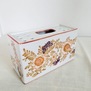 May include: White rectangular ceramic tissue box with a floral and butterfly design in shades of orange, brown, and purple. The box has a rectangular opening on top and a series of small holes. The edges are trimmed in a reddish-brown color.
