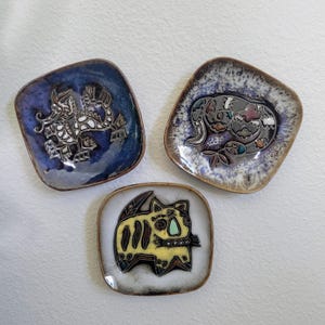 May include: Three square ceramic dishes with rounded corners. The top left dish is deep blue with a white dragon motif. The top right dish features a mottled white and blue glaze with a dark, stylized animal figure. The bottom dish is white with a yellow cat.