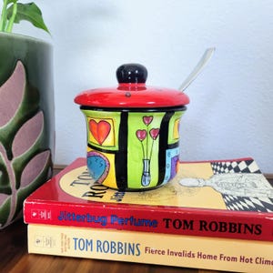 May include: A ceramic jar with a bright red lid and a black knob, featuring a vibrant lime green body decorated with colorful hand-painted squares containing hearts and floral motifs. A silver metal spoon rests inside the jar, which is placed on books.