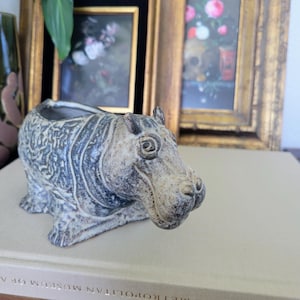 May include: A ceramic hippo planter in shades of gray and beige. The hippo has a detailed textured design and a wide opening for plants. The planter is sitting on a book with framed art in the background.