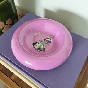 May include: A round, light pink glass dish holding several pieces of jewelry. The jewelry includes a pair of floral earrings, a pair of small silver earrings, and a pair of blue and silver earrings. The dish is sitting on a purple surface.
