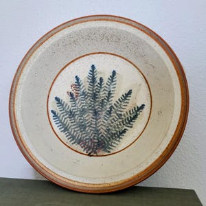 May include: A round ceramic plate with a fern design. The plate features a cream-colored center with a blue and green fern illustration. The rim is a speckled beige with a brown border. The plate is a decorative piece for serving food.