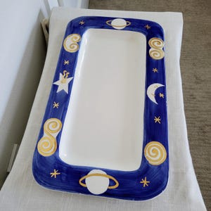 May include: Rectangular white serving platter with a deep blue border. The border is decorated with gold and white celestial designs, including stars, planets, and swirls. The platter is resting on a white cloth.