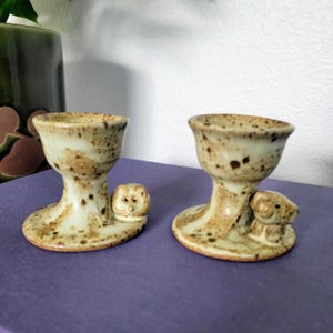 May include: Two ceramic egg cups with a speckled, light brown glaze. Each cup has a pedestal base and a small, sculpted dog figurine. The egg cups are displayed on a purple surface.
