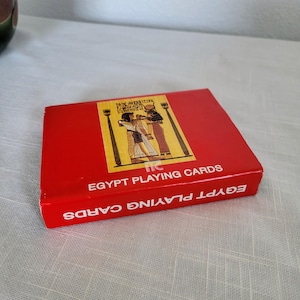 May include: A white ceramic mug with a black handle and the text "But First, Coffee" in a bold, black font. The mug is set against a blurred background, suggesting a cozy setting.