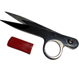 May include: A pair of small, silver scissors with black blades and a finger hole. A red protective cover is next to the scissors. The scissors are designed for detailed cutting tasks.