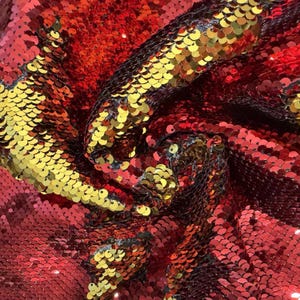 May include: Close-up of a fabric with red, gold, and black sequins. The sequins are arranged in a scale-like pattern, creating a shimmering effect. The fabric is folded and draped, showing the texture and depth of the material.