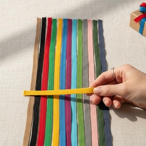 Puede incluir: Una selección de cintas de colores dispuestas verticalmente, con una mano sosteniendo una cinta amarilla horizontalmente. En el fondo, una pequeña caja de regalo envuelta en papel marrón y atada con cinta azul, roja y azul.