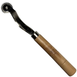 May include: A sewing tracing wheel featuring a circular, serrated metal gear attached to a curved, silver-toned metal shank. The tool includes a cylindrical, light brown wooden handle with a smooth finish and a silver metal ferrule at the base.