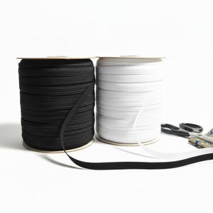 May include: Two spools of elastic ribbon, one black and one white, sit side-by-side against a white background. A pair of scissors and a small piece of patterned fabric are to the right of the spools. The black elastic ribbon is unspooled.