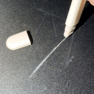 May include: A white stylus with a black tip and a white cap on a black surface. The stylus is creating a white line on the surface. The image shows the stylus and cap.