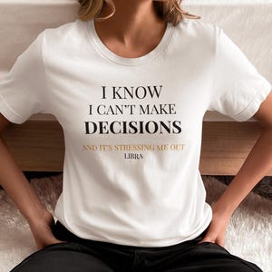 Puede incluir: Camiseta blanca con el texto "I KNOW I CAN'T MAKE DECISIONS AND IT'S STRESSING ME OUT LIBRA" en letras negras y doradas. La camiseta tiene cuello redondo y mangas cortas. La persona lleva pantalones negros.