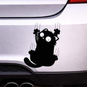 Puede incluir: Adhesivo de gato negro aferrado a la superficie de un coche. El gato de dibujos animados tiene ojos grandes y araña con sus garras, creando un efecto juguetón y humorístico. El adhesivo está diseñado para parecer que el gato está trepando.