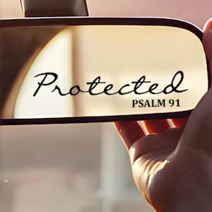 Op de afbeelding: Een achteruitkijkspiegel van een auto met het woord "Protected" in cursief schrift en "PSALM 91" eronder. De spiegel heeft een bruine lijst en wordt door een hand vastgehouden, met een vage achtergrond.