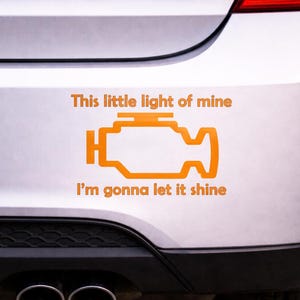 May include: Orange sticker on a car bumper with an engine light symbol and the words "This little light of mine, I'm gonna let it shine."