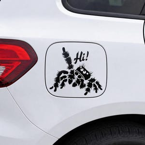 Puede incluir: Una calcoman&iacute;a negra de ara&ntilde;a con la palabra "Hi!" sobre un fondo blanco, adherida a la tapa del combustible de un coche blanco. La ara&ntilde;a tiene un dise&ntilde;o de dibujos animados con ojos grandes y patas peludas. Un divertido accesorio decorativo para el coche.