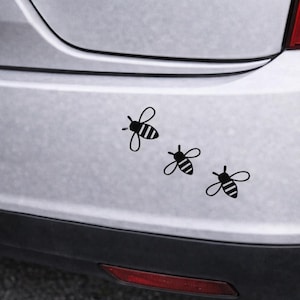 May include: Three black bee decals are arranged diagonally on a silver car bumper. Each bee has striped abdomens and delicate wings, creating a playful and whimsical design. The decals add a touch of nature-inspired charm.