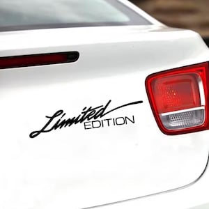 以下が含まれることがあります： 白い車の後部に貼られた黒い「Limited Edition」ステッカー。テキストは筆記体で、「Limited」が「EDITION」の上にあります。車の赤いテールライトが画像右側に表示されています。