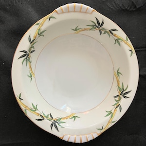 May include: White ceramic serving bowl with a bamboo design. The bowl features a gold rim and handles, with green and black bamboo leaves and yellow bamboo stalks. The bowl has a round interior and a square exterior.