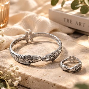 May include: A silver-toned metal cuff bracelet and a matching open ring, both featuring a detailed fish design with textured scales and fins. The bracelet has two fish heads meeting at the center, while the ring features a similar dual-fish motif.