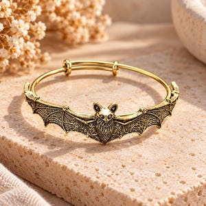 Op de afbeelding: Een goudkleurige metalen armband met een centraal vleermuismotief met uitgestrekte vleugels. Het vleermuisontwerp heeft gedetailleerde, getextureerde gravures op de vleugels en het lichaam, en de armband heeft een dunne, verstelbare ringband.