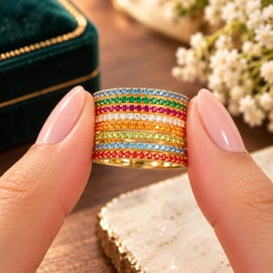May include: A gold band ring featuring multiple horizontal rows of small, round, multi-colored gemstones in shades of blue, green, pink, white, orange, yellow, and red, arranged in a vibrant, stacked, rainbow-inspired pattern.