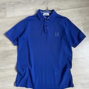 Puede incluir: Un polo azul brillante con mangas cortas, cuello y tapeta de tres botones. La camisa tiene un pequeño parche cuadrado con el logo en el pecho y está colgada de una percha metálica.