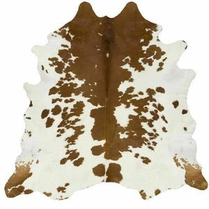 Puede incluir: Una alfombra de piel de vaca con un patrón natural de manchas marrones y blancas. La alfombra tiene una forma irregular, con una gran sección marrón en el centro y bordes blancos. La piel es probablemente de una vaca.