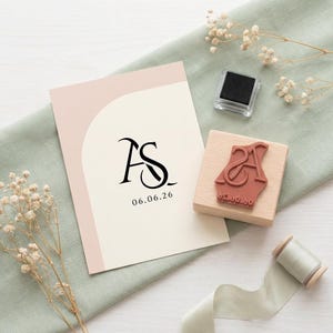 May include: A wedding invitation featuring the initials "AS" and the date "06.06.26" in black. A wooden stamp with the same initials and date is positioned beside the invitation, alongside a black ink pad and a ribbon spool. The backdrop is a pale green cloth.