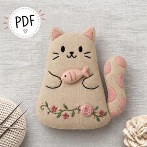 May include: A beige fabric cat plushie featuring embroidered black facial features, a pink fish held in its paws, and a pink polka-dot tail. The lower body displays an embroidered green vine with pink flowers. A circular white tag reads PDF.