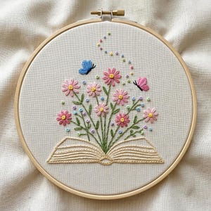 May include: An embroidery hoop containing a piece of off-white fabric with a hand-stitched design of an open book. Pink daisies with green stems emerge from the pages, accompanied by a blue butterfly, a pink butterfly, and small colorful dots.
