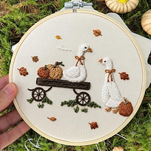 May include: A circular embroidery hoop containing a cream-colored fabric with a hand-stitched design of two white geese, one pulling a dark brown wooden wagon filled with two orange pumpkins, and another goose standing beside a third pumpkin.