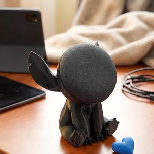 May include: A black figurine of a cartoon character with large ears and a round head, sitting on a wooden surface. A small blue heart-shaped object is next to the figurine. A tablet and a cable are also visible in the background.
