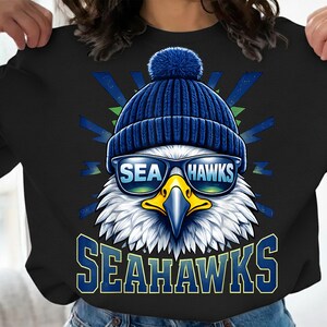 May include: Black sweatshirt with a graphic of an eagle wearing a blue beanie and sunglasses that read "SEA HAWKS". The eagle has a yellow beak, and the word "SEAHAWKS" is printed in blue below.
