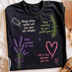 以下が含まれることがあります： カラフルなグラフィックとテキストがプリントされた黒いTシャツ。デザインには、Always throw spilled salt over your left shoulderというテキストが添えられた塩入れ、Keep rosemary by your garden gateというテキストが添えられたローズマリーの小枝、Plant lavender, for luckというテキストが添えられたラベンダーが含まれています。ピンクのハートにはFall in love whenever you canというテキストが添えられています。