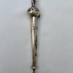 Może przedstawiać: Srebrny, zabytkowy otwieracz do listów o szczegółowym wzornictwie. Mała figurka siedzi na sferycznym uchwycie, a wydłużone ostrze ma elementy dekoracyjne. Otwieracz ma około 23 cm długości.