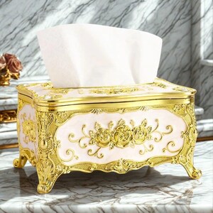 Peut inclure: Un élégant distributeur de mouchoirs avec un cadre et des pieds dorés. Le centre est rose pâle avec des motifs floraux dorés. Des mouchoirs blancs dépassent du haut. Un objet décoratif pour la maison.
