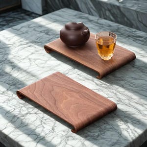 Może przedstawiać: Dwie drewniane tace do serwowania z zakrzywionymi krawędziami. Jedna taca zawiera mały brązowy imbryk. Druga taca zawiera przezroczysty szklany kubek ze złotym płynem. Tace wykonane są z ciemnego drewna z widocznym usłojeniem.