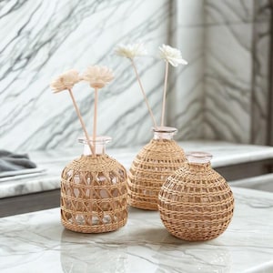 Pode incluir: Três vasos de vidro transparente com exteriores de rattan tecido. Dois vasos contêm flores decorativas brancas e bege. Os vasos estão dispostos sobre uma superfície de mármore, criando uma estética natural e elegante.